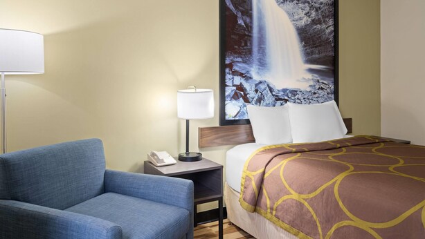 A hotel room with a bed, a nightstand, a lamp, a chair, and a large waterfall painting on the wall. - Super 8 by Wyndham Hagerstown I-70 (Hagerstown)
