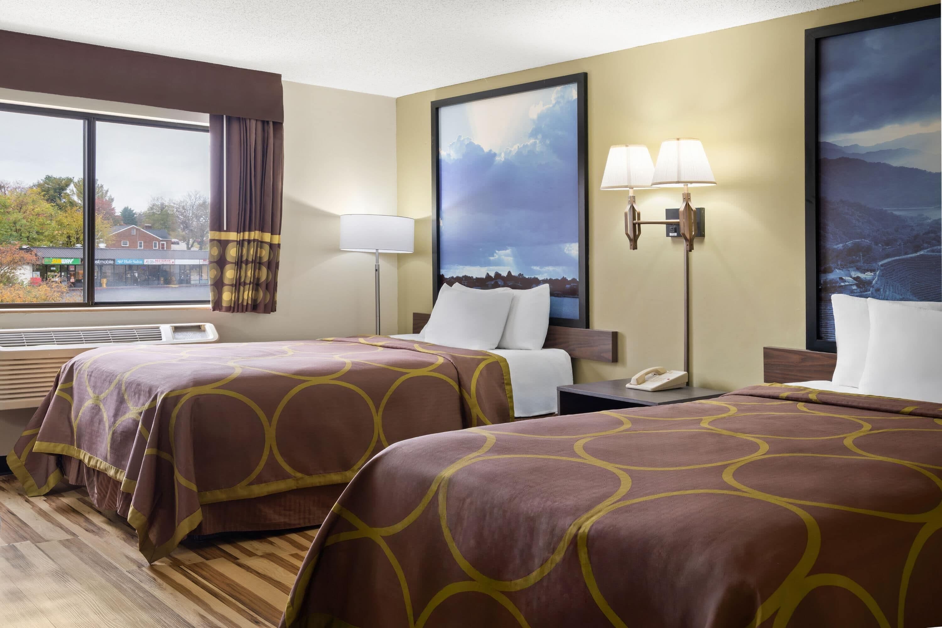 Une chambre d’hôtel avec deux lits, une grande fenêtre avec des rideaux, un téléphone fixé au mur et un tableau représentant un paysage.