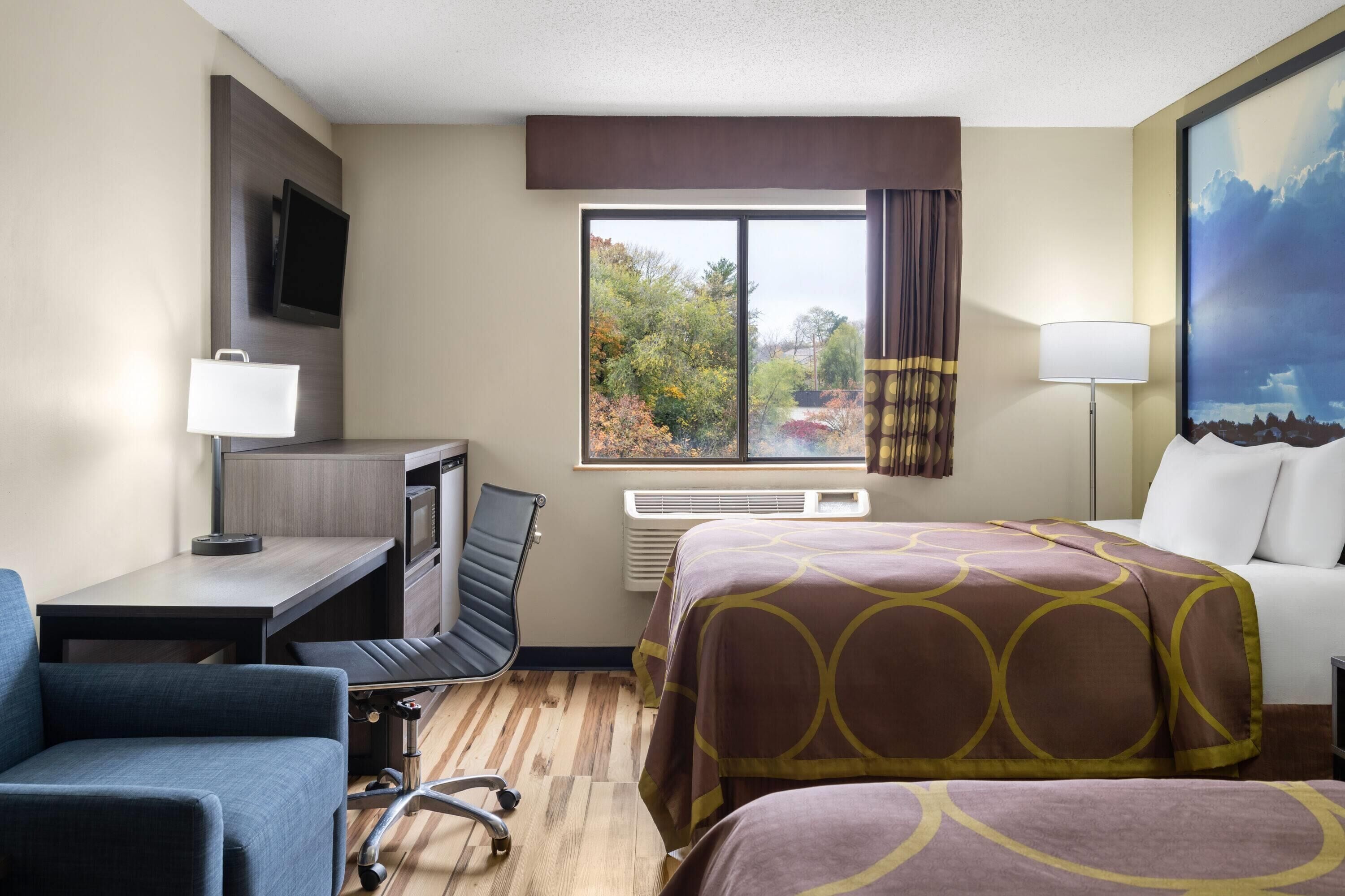 Une chambre d’hôtel équipée d’un lit, d’un bureau, d’une chaise, d’une télévision et d’une fenêtre donnant sur la verdure.