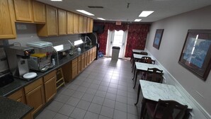 Una cocina con armarios de madera, una encimera con un tostador, un fregadero y una ventana con cortinas rojas. Hay mesas y sillas en la zona de comedor.