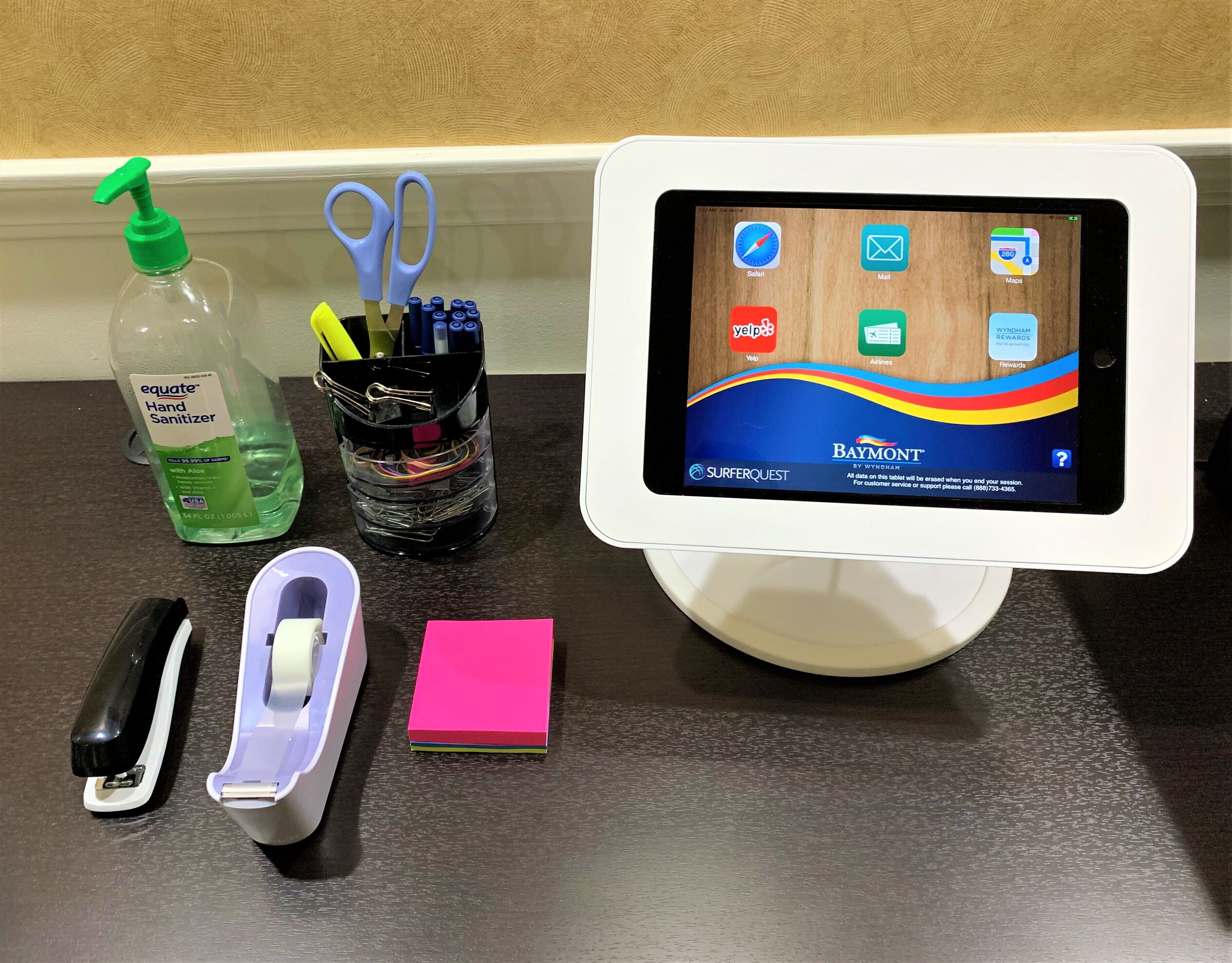 A tablet displaying a rewards program interface, a bottle of hand sanitizer, a container with pens and scissors, a stapler, and a sticky note on a counter.
