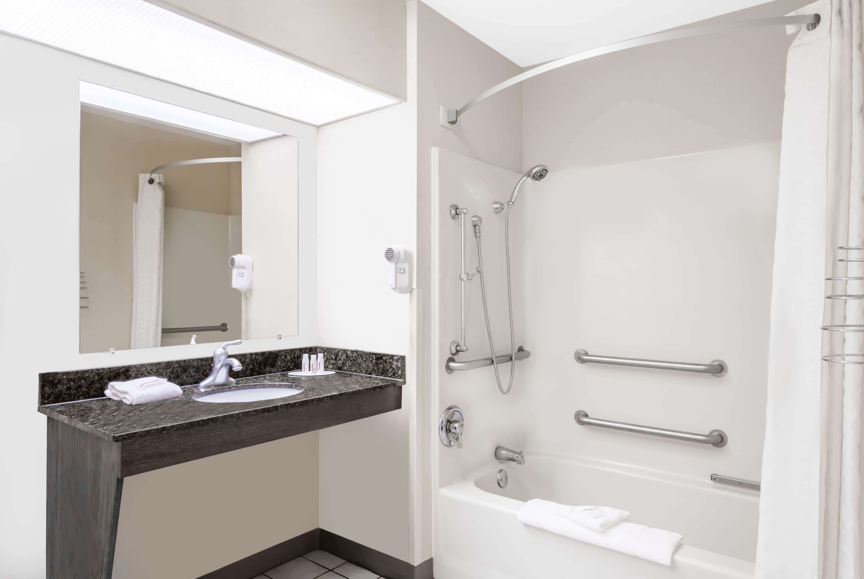 Un baño moderno con lavamanos de granito, un espejo grande, una bañera con barras de apoyo y una ducha.