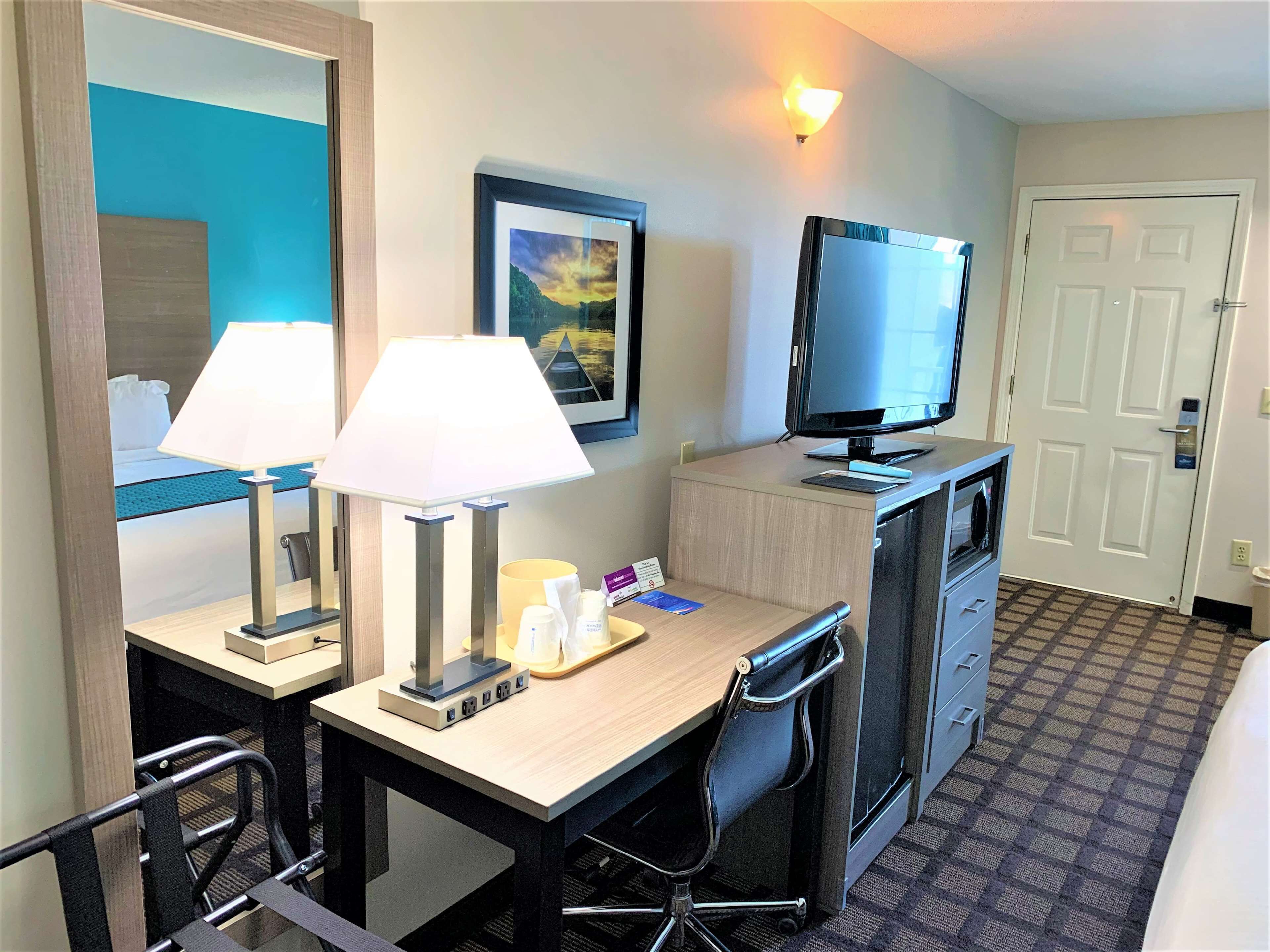 A hotel room with a desk, two lamps, a television, a chair, a dresser, and a door.