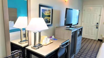 A hotel room with a desk, two lamps, a television, a chair, a dresser, and a door.