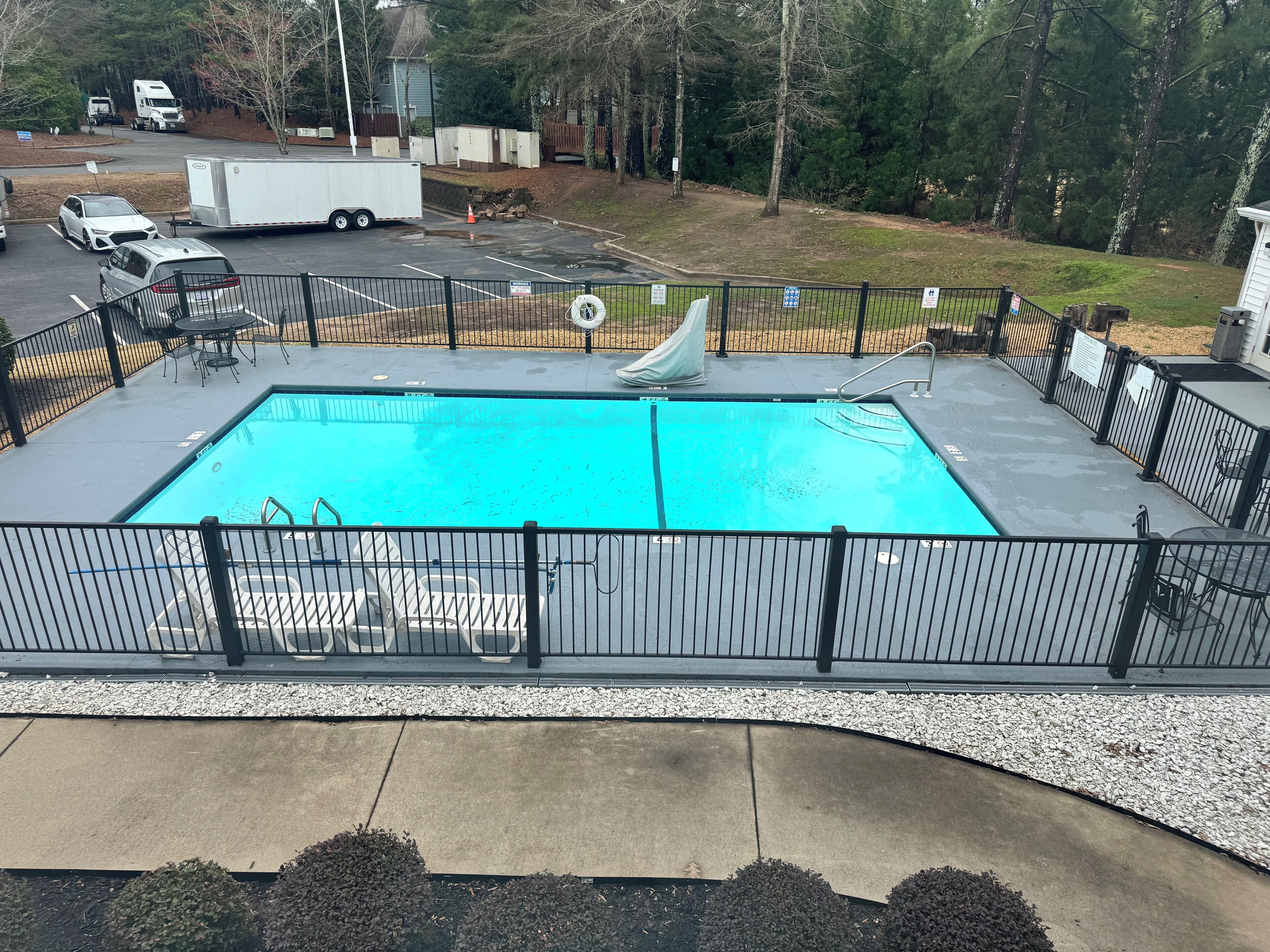 A rectangular swimming pool with a slide, surrounded by a black metal fence, and a concrete deck with lounge chairs.