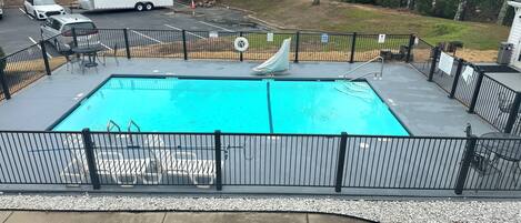 A rectangular swimming pool with a slide, surrounded by a black metal fence, and a concrete deck with lounge chairs.