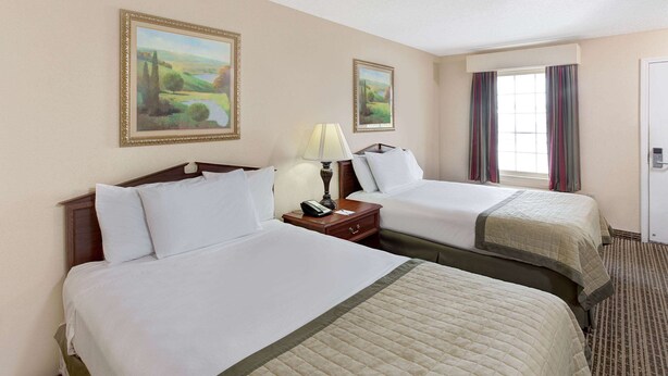 A hotel room with two beds, a nightstand with a lamp, and two framed landscape paintings on the wall. - Baymont by Wyndham Sanford (Sanford)