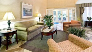 A spacious living room with a sofa, armchairs, a coffee table, and a potted plant. There is a door leading to a balcony with a view of the outdoors. - Baymont by Wyndham Sanford (Sanford)