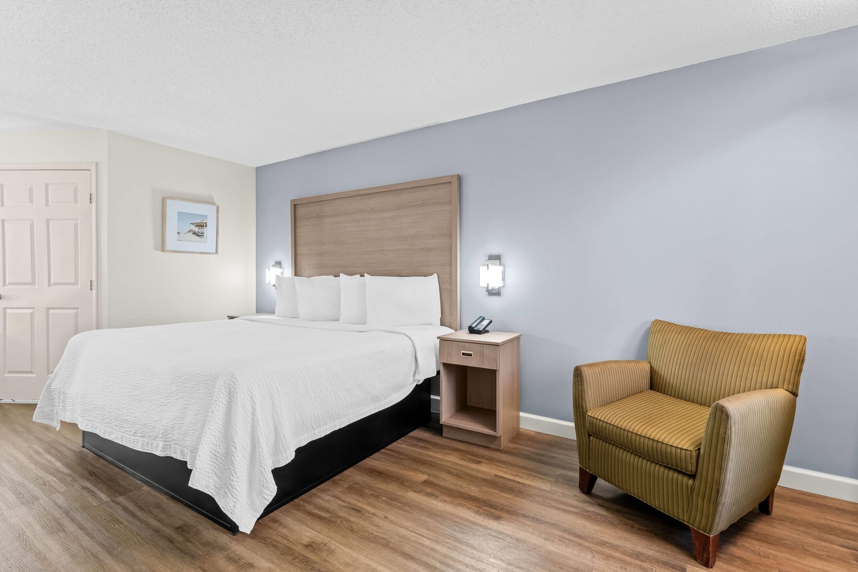 A hotel room with a bed, bedside table, chair, and a painting on the wall.