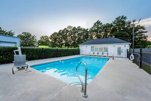 Una piscina con una escalera metálica, rodeada por una valla, con un edificio y árboles al fondo.
