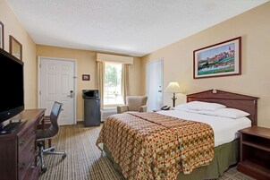 Una habitación de hotel con una cama, un escritorio con televisor, una silla, una lámpara y un cuadro en la pared.