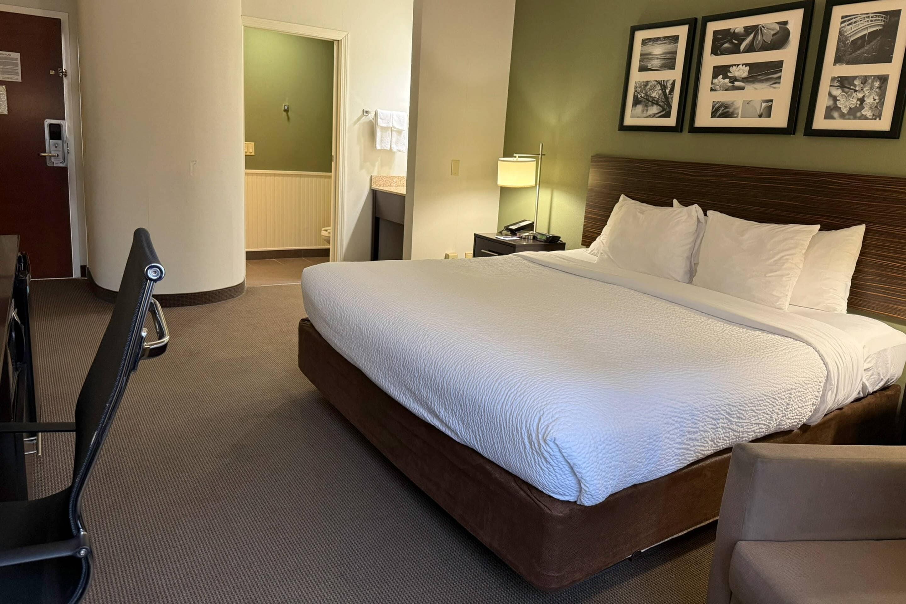 A hotel room with a large bed, a nightstand with a lamp, a chair, and framed pictures on the wall.