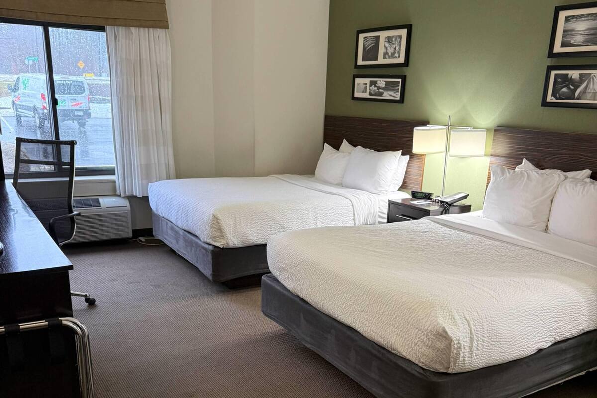 A hotel room with two beds, a desk, and a chair. There are framed pictures on the wall and a window with curtains.