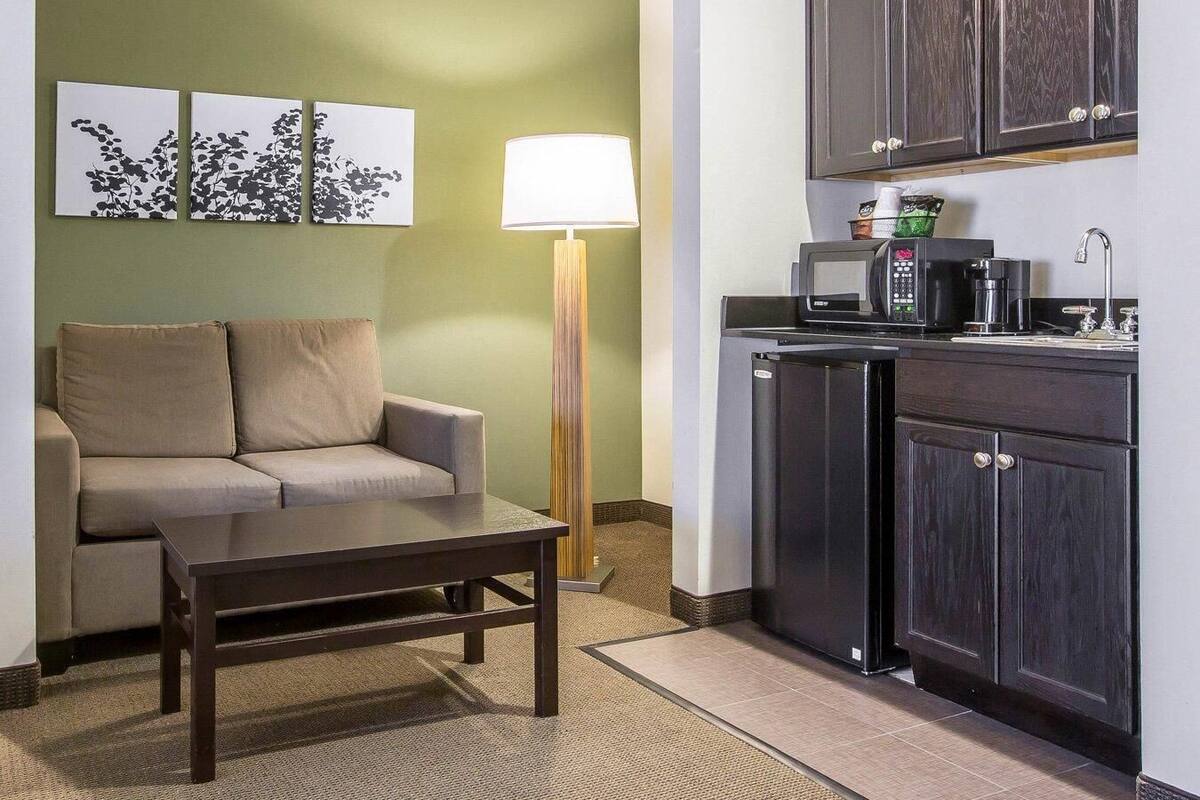 A compact hotel room with a sofa, a small table, a floor lamp, a kitchenette with a microwave, coffee maker, and cabinets.