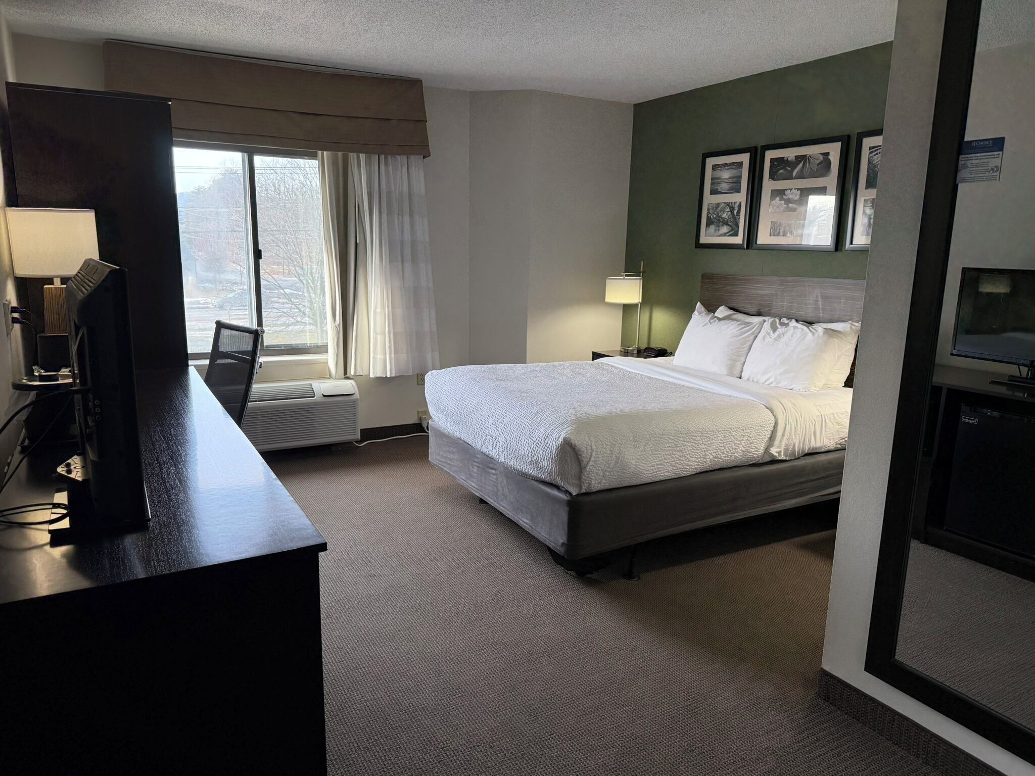 A hotel room with a bed, a desk with a computer, a window with curtains, and framed pictures on the wall.