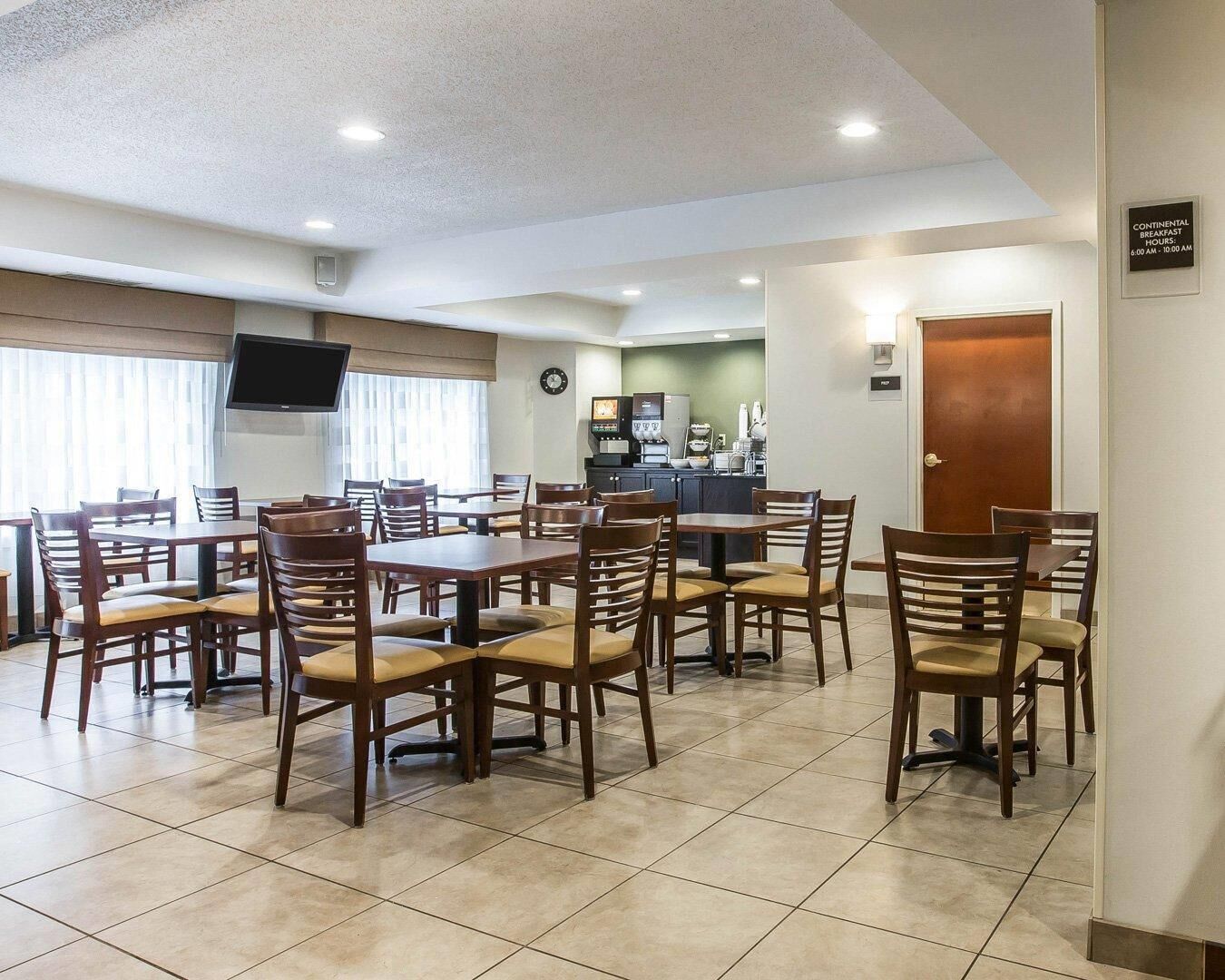 A hotel dining area with multiple tables and chairs, a kitchen in the background, and a sign indicating continental breakfast hours.