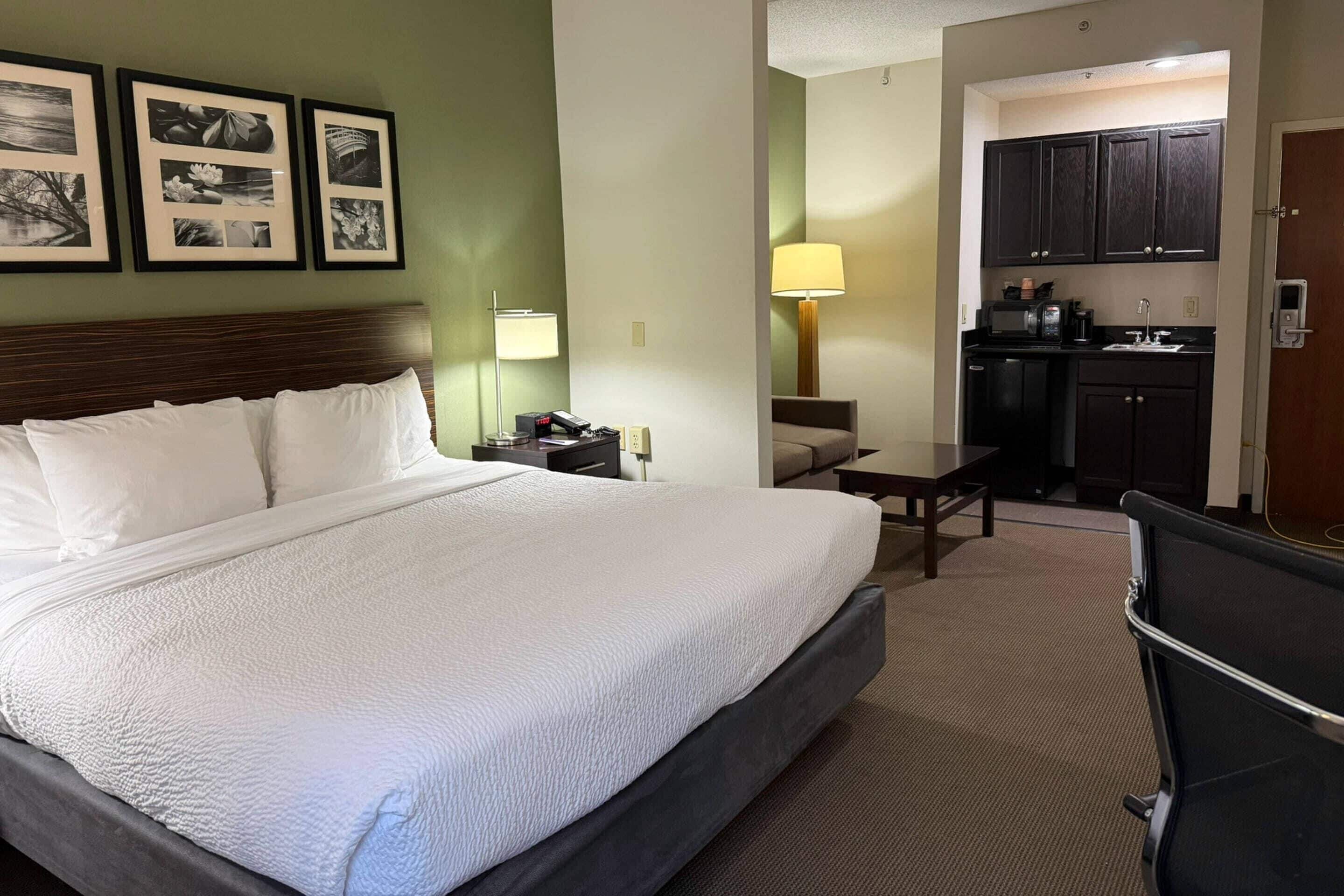 A hotel room with a large bed, a desk, a chair, a kitchenette, and framed pictures on the wall.