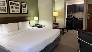 A hotel room with a large bed, a desk, a chair, a kitchenette, and framed pictures on the wall.