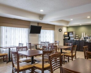 A hotel dining area with multiple tables and chairs, a television mounted on the wall, a clock, and a kitchenette with a coffee machine.