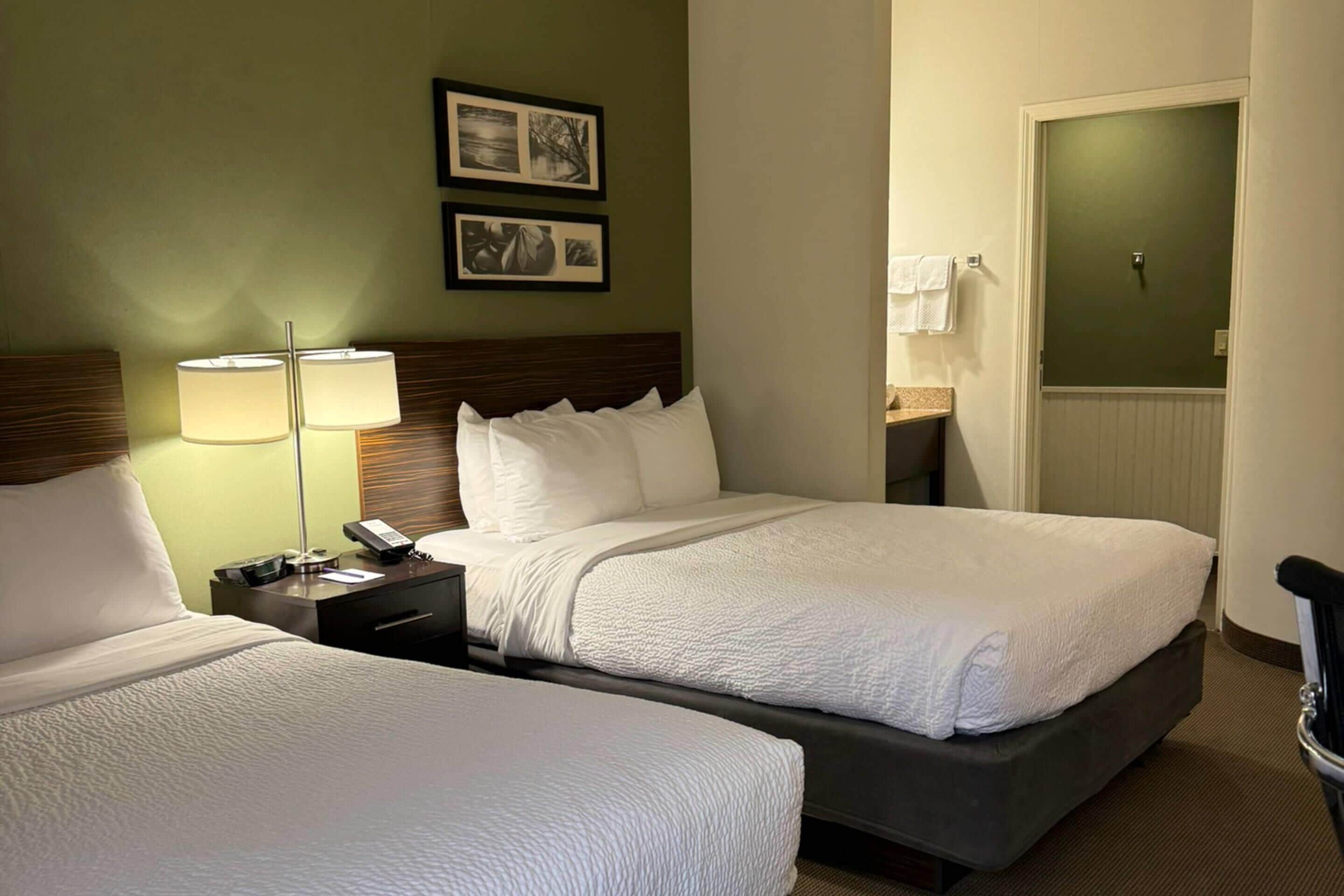 Habitación de hotel con dos camas, una mesita de noche con lámpara y puerta al baño.
