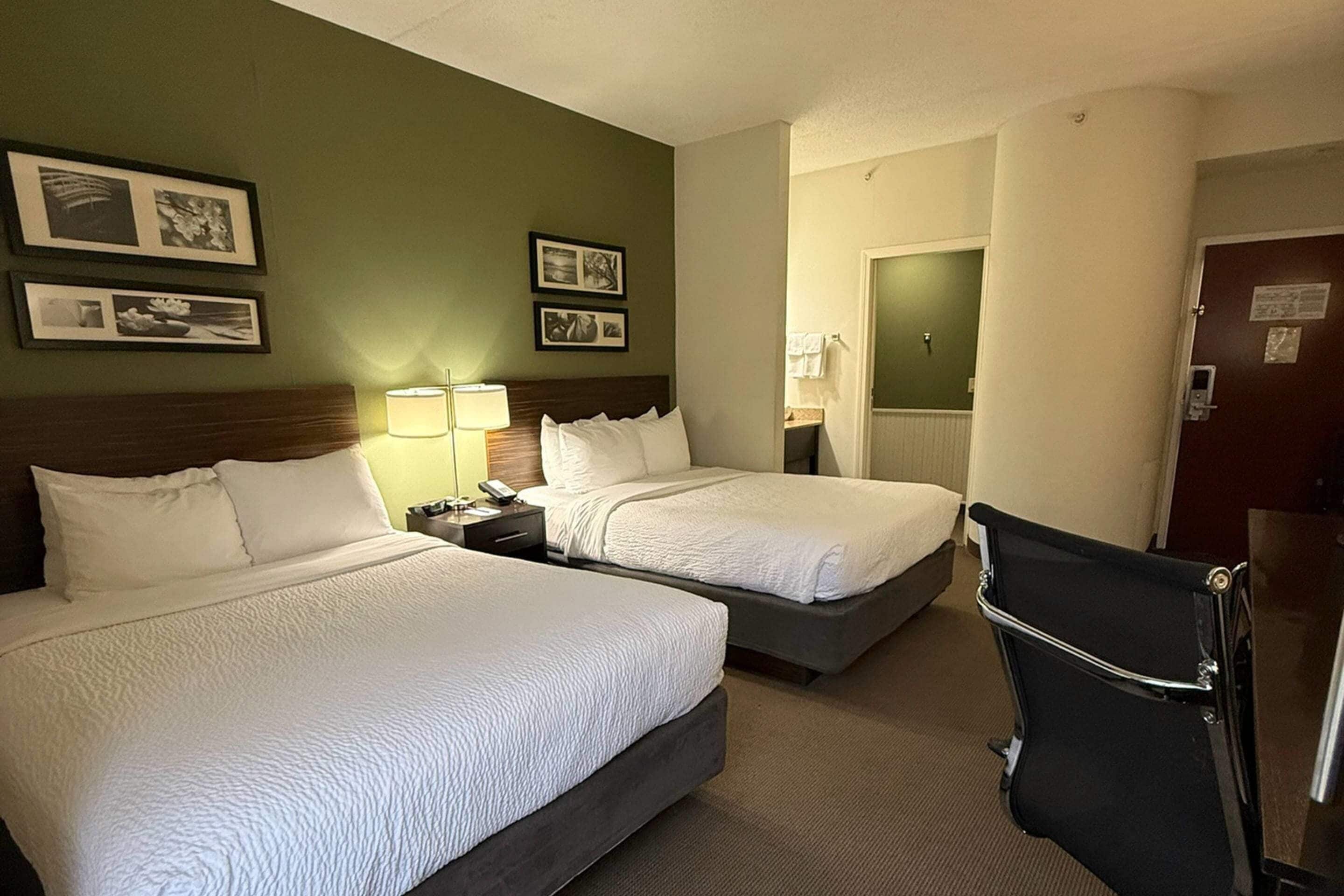 A hotel room with two beds, a desk, a chair, and a bathroom visible through an open door.