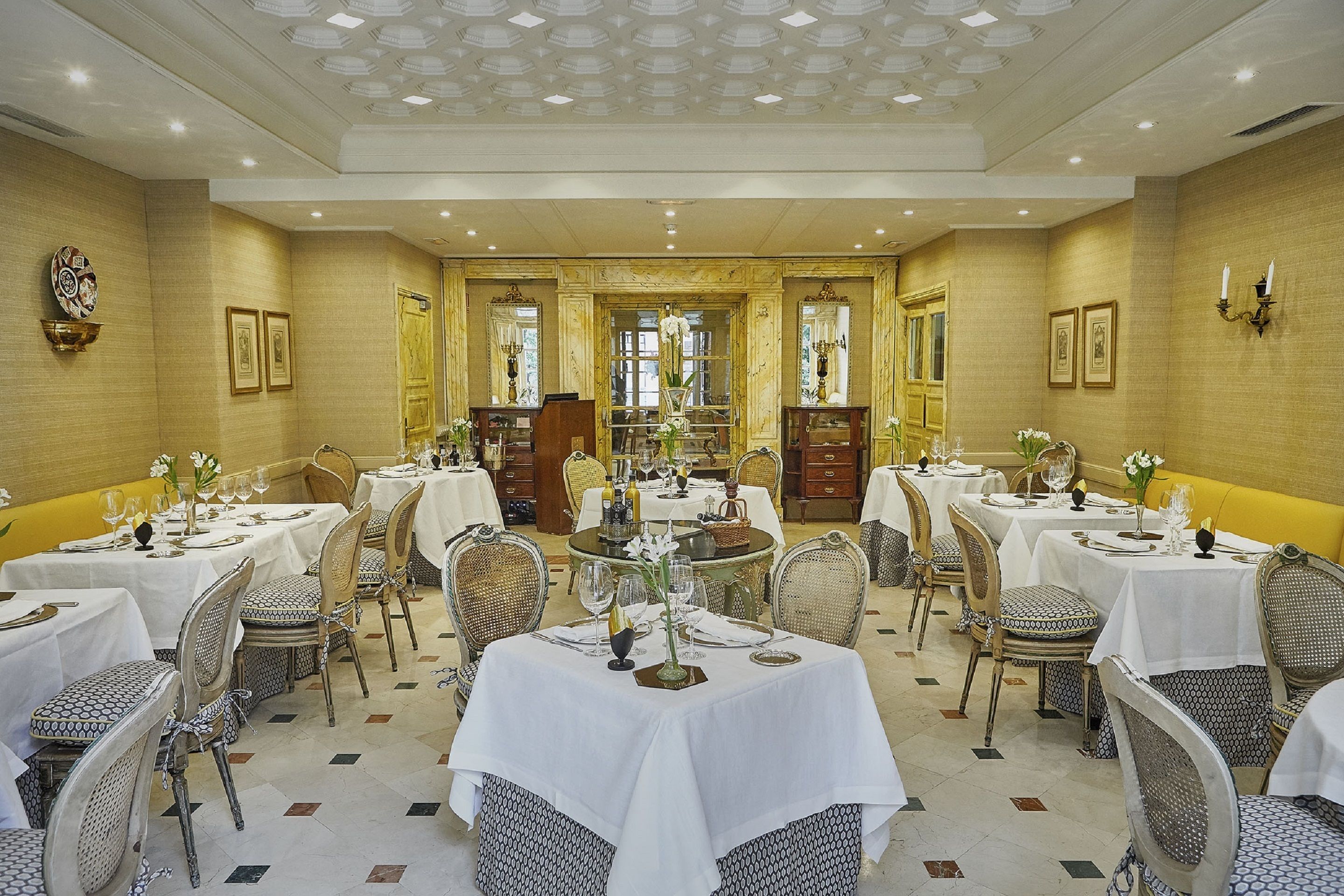 A formal dining room with multiple tables and chairs, each set with white tablecloths and silverware.
