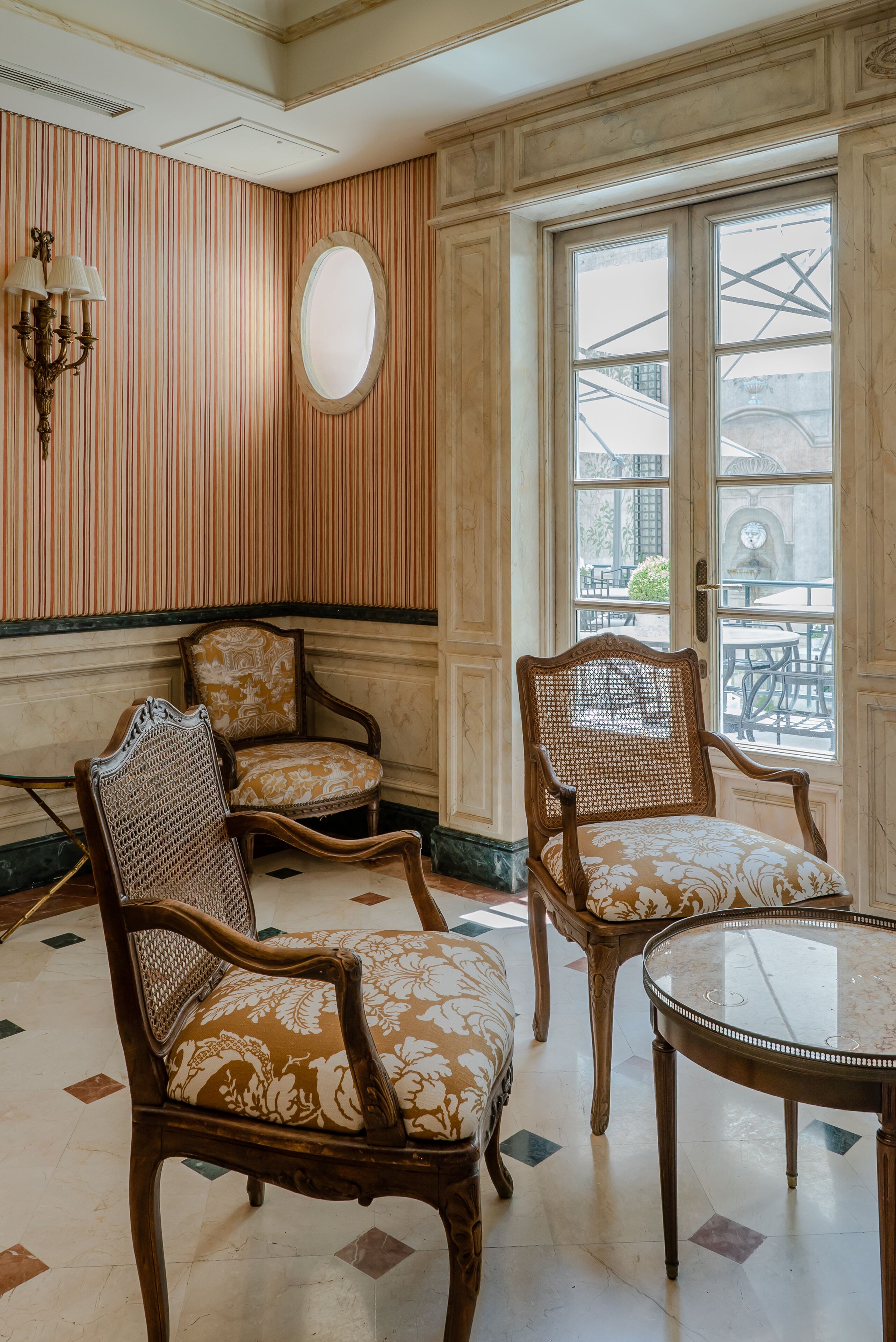 A room with two ornate wooden chairs with patterned cushions, a round table, and a large window with a view of the outside.