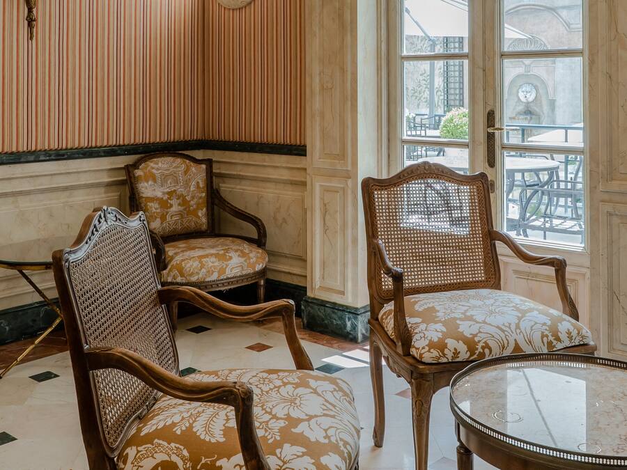 A room with two ornate wooden chairs with patterned cushions, a round table, and a large window with a view of the outside.