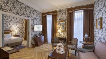 Habitación de hotel con cama, sofá, mesa de comedor y dos sillas. Hay un televisor en la pared y una mesita de noche con lámpara.