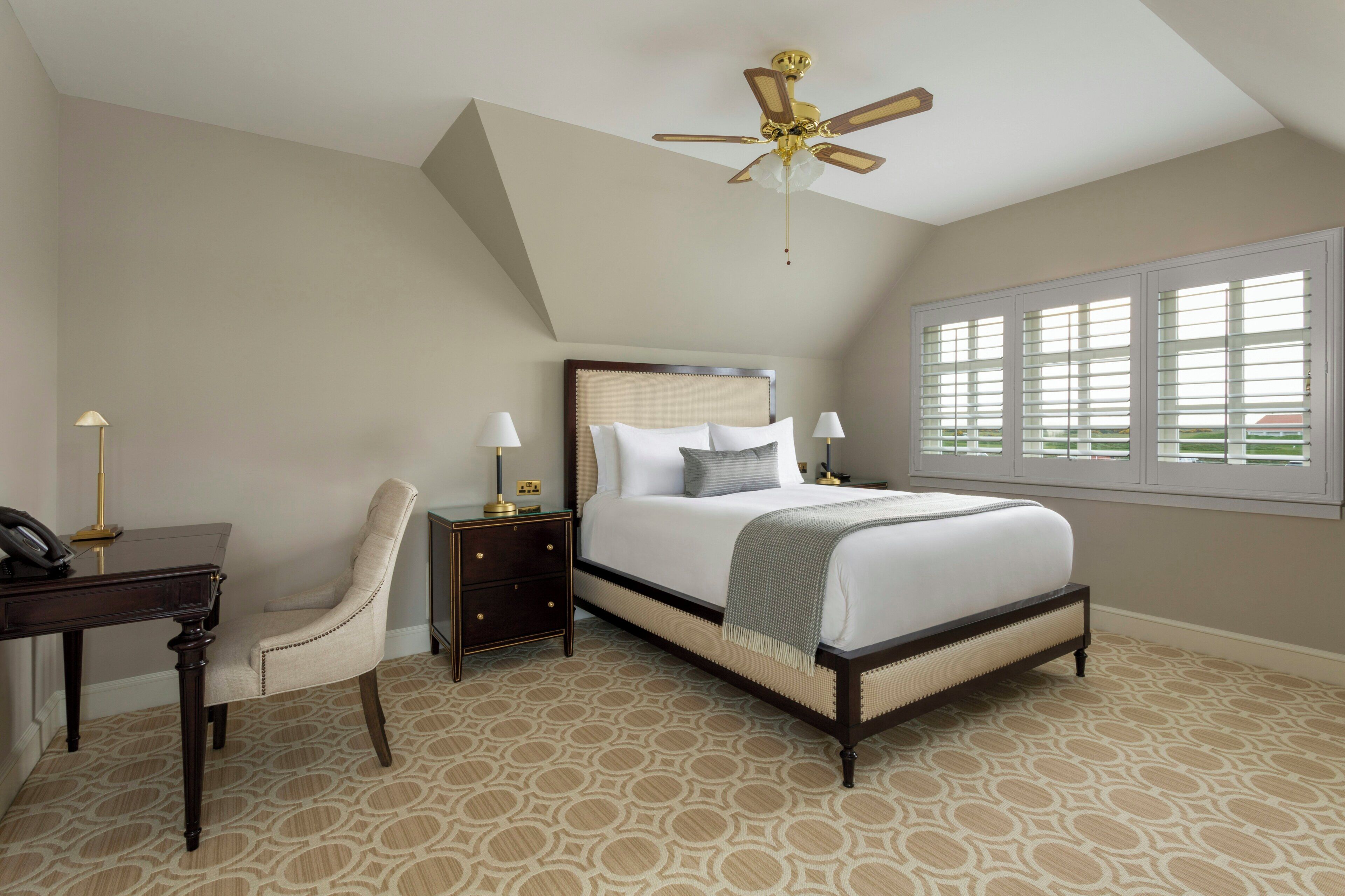 A bedroom with a bed, a desk, a chair, a nightstand, a ceiling fan, and a window with blinds.
