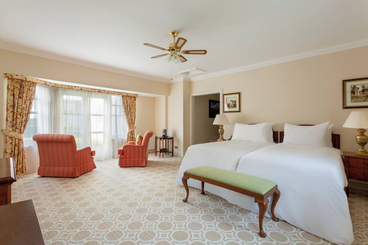 A hotel room with a large bed, two armchairs, a desk, a ceiling fan, and a patterned carpet.