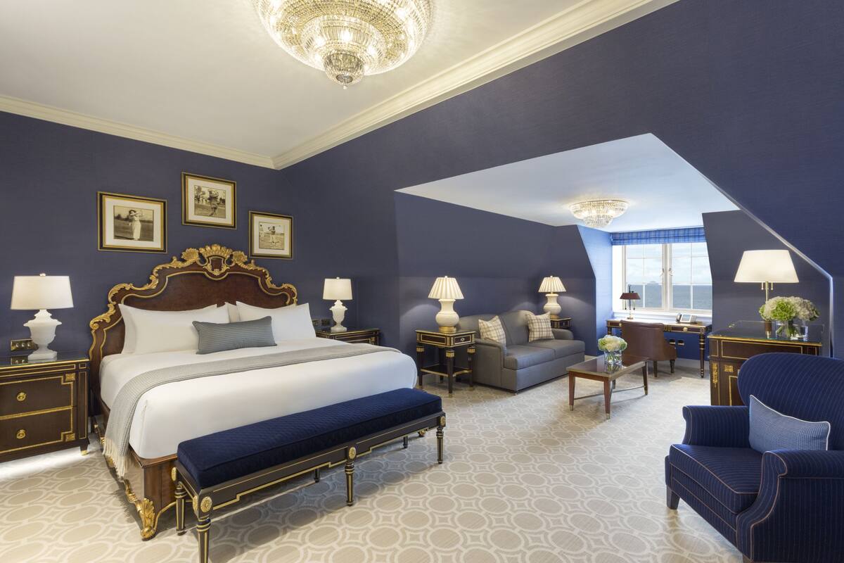 A spacious bedroom with a large bed, a sitting area with a sofa and armchairs, and a desk with a lamp.