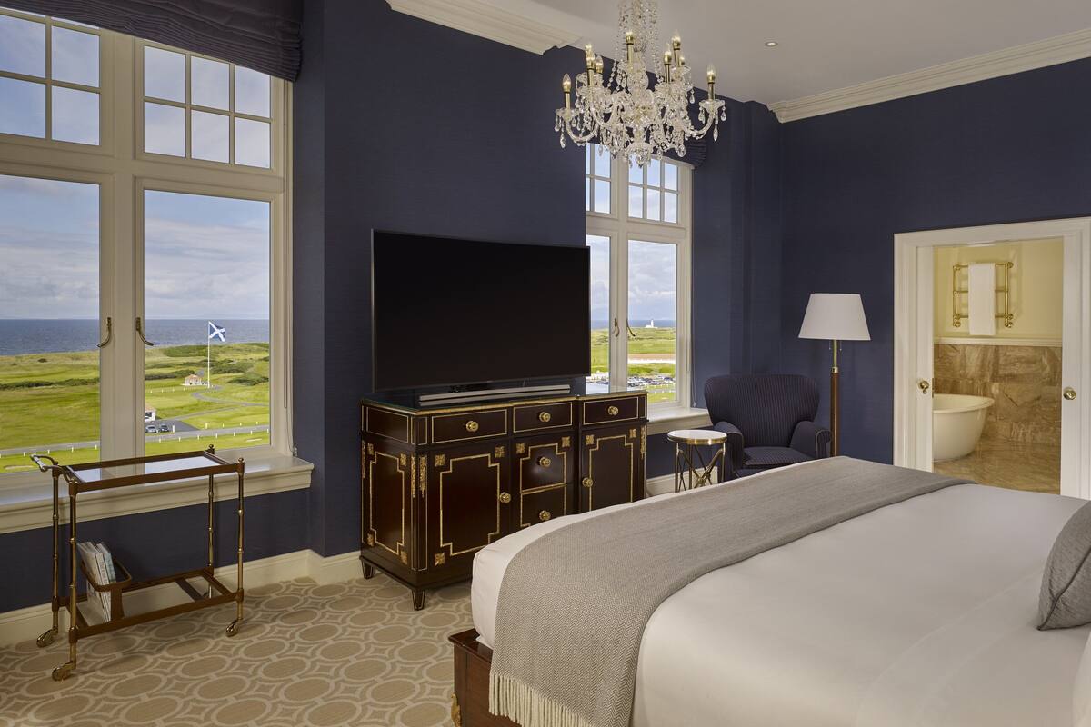 A bedroom with a large bed, a TV on a dark wooden dresser, a chandelier, and a view of the sea through large windows.