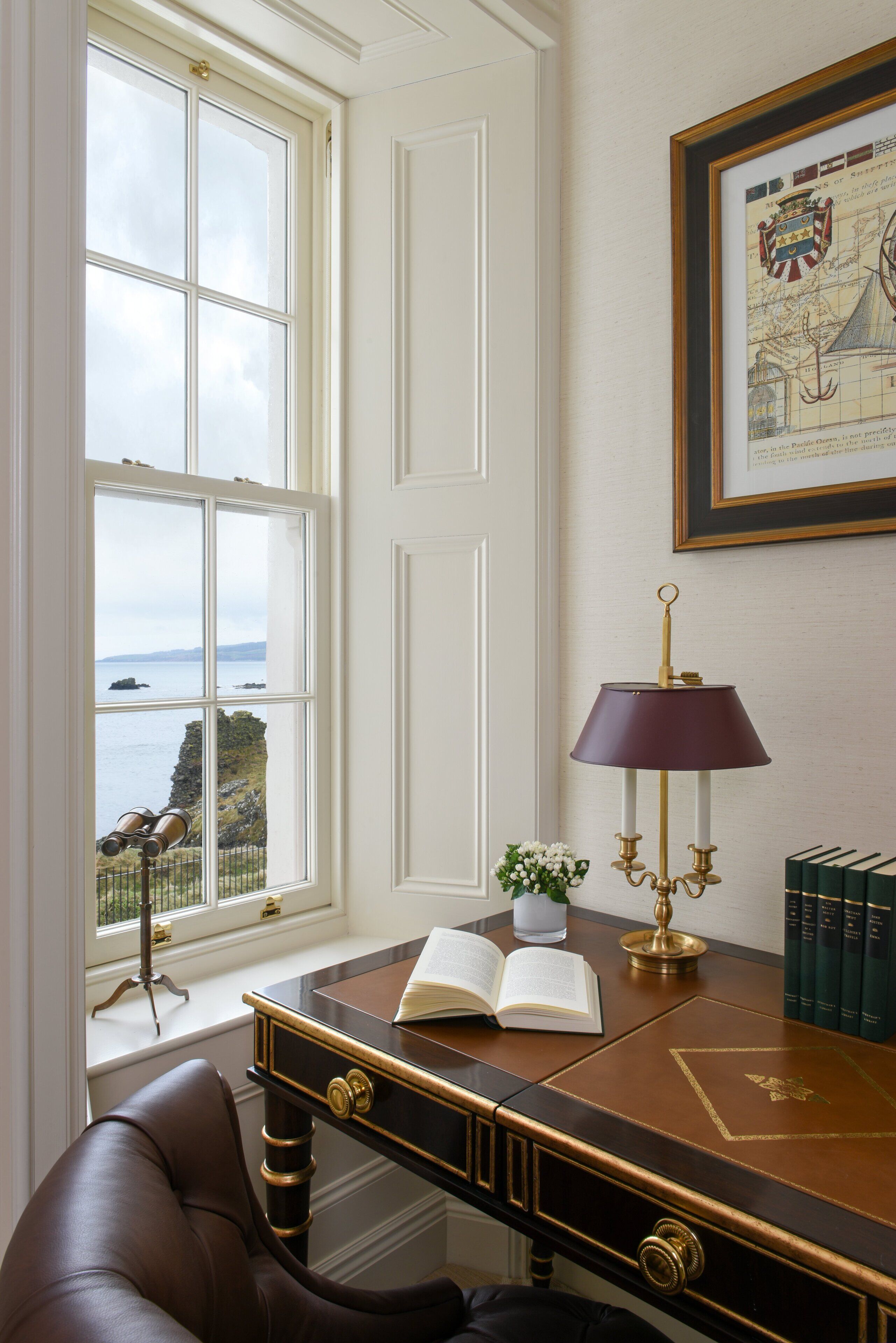 A study room with a desk, books, a lamp, and a window with a view of a coastal landscape.