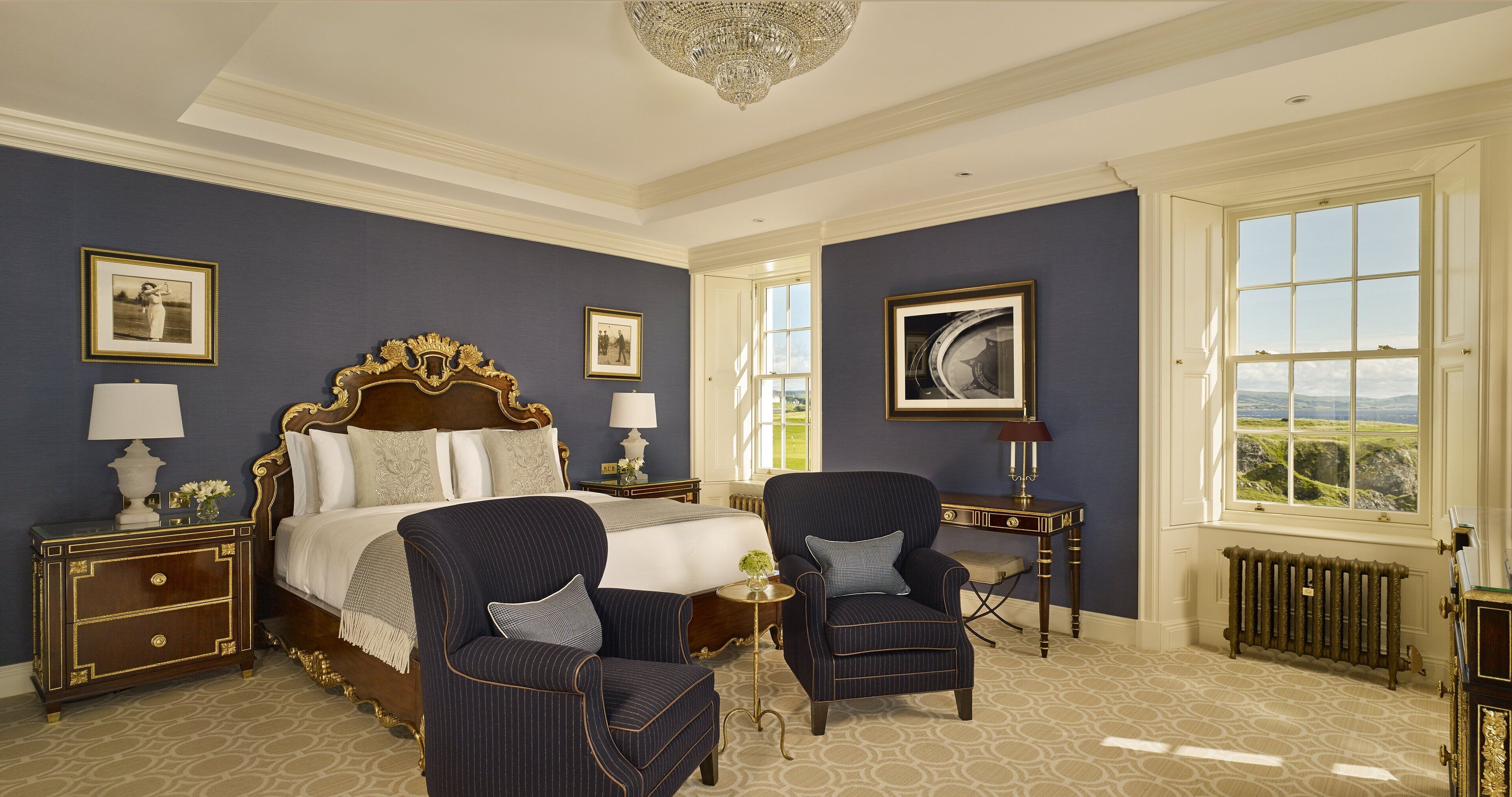Une chambre à coucher avec un grand lit, deux fauteuils, une table de chevet, une commode et une fenêtre donnant sur un beau paysage.