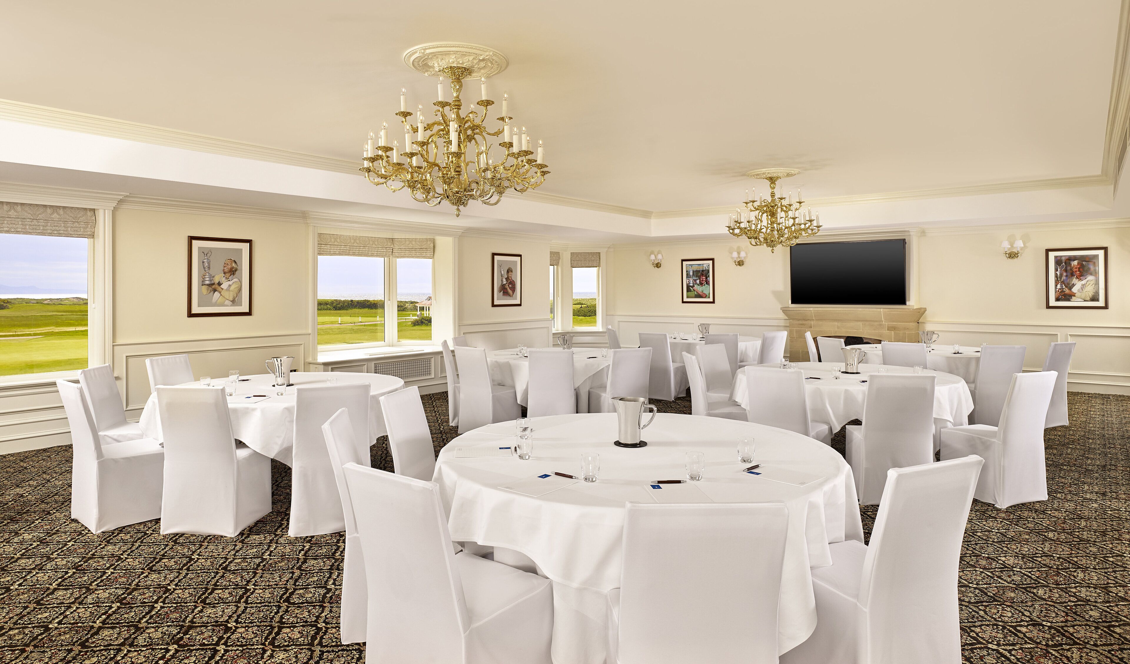 Un salón de conferencias con mesas redondas, sillas blancas, candelabros y ventanas amplias con vista a un campo de golf.