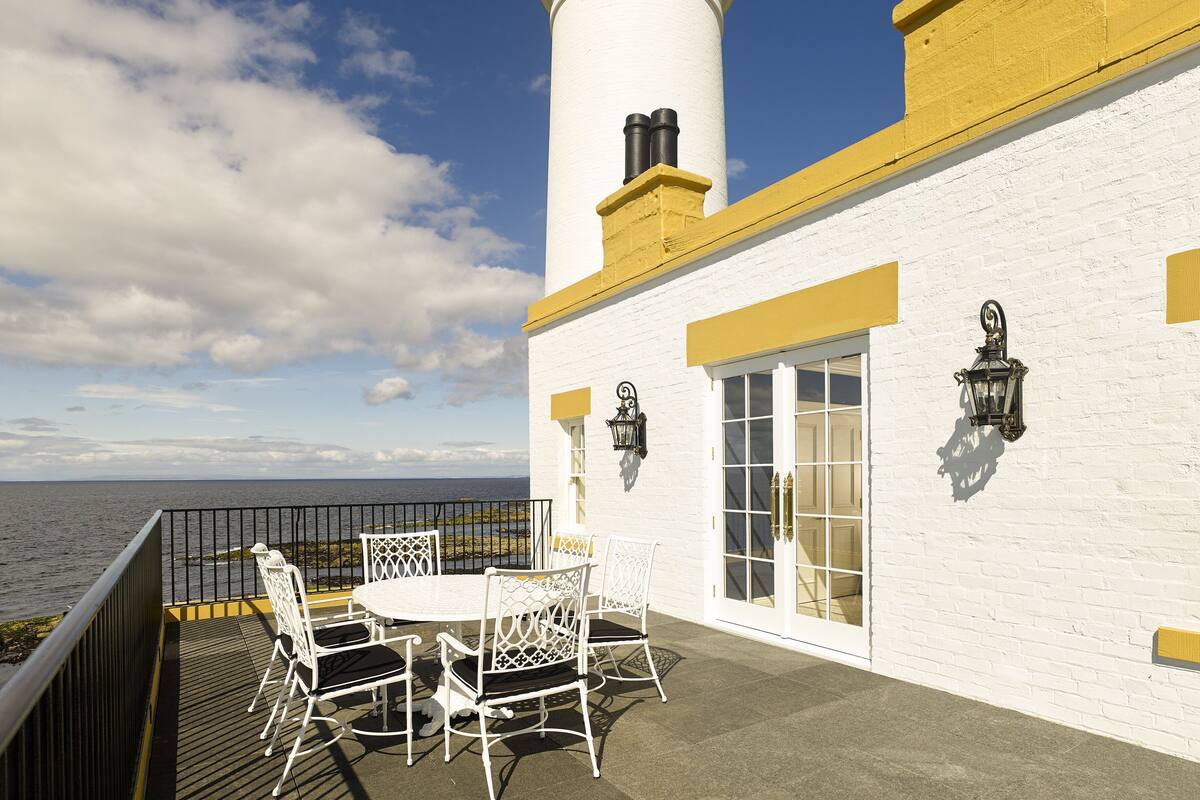 A white building with a lighthouse, a table with chairs, and a view of the sea.