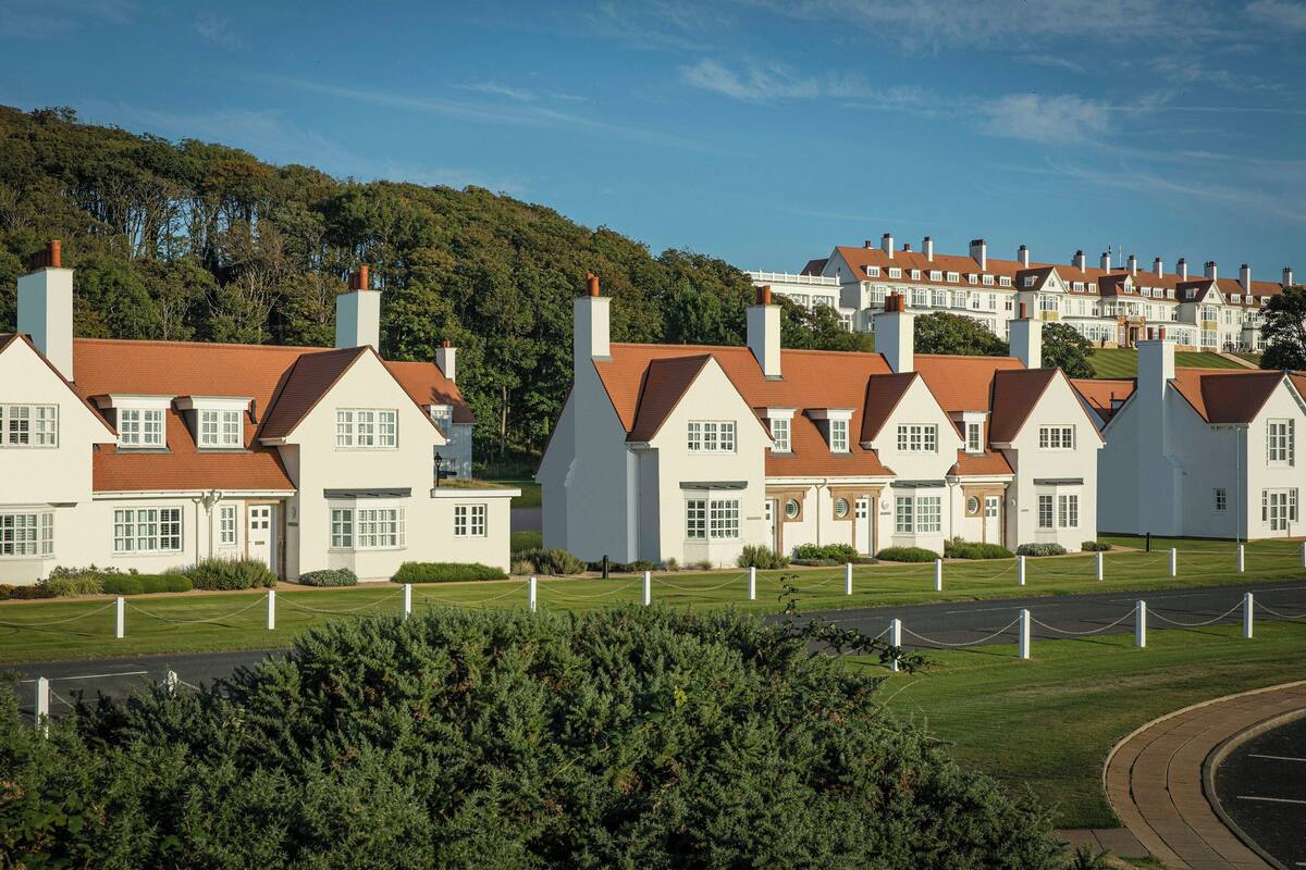 A row of white houses with red roofs, surrounded by greenery and a hilly backdrop.