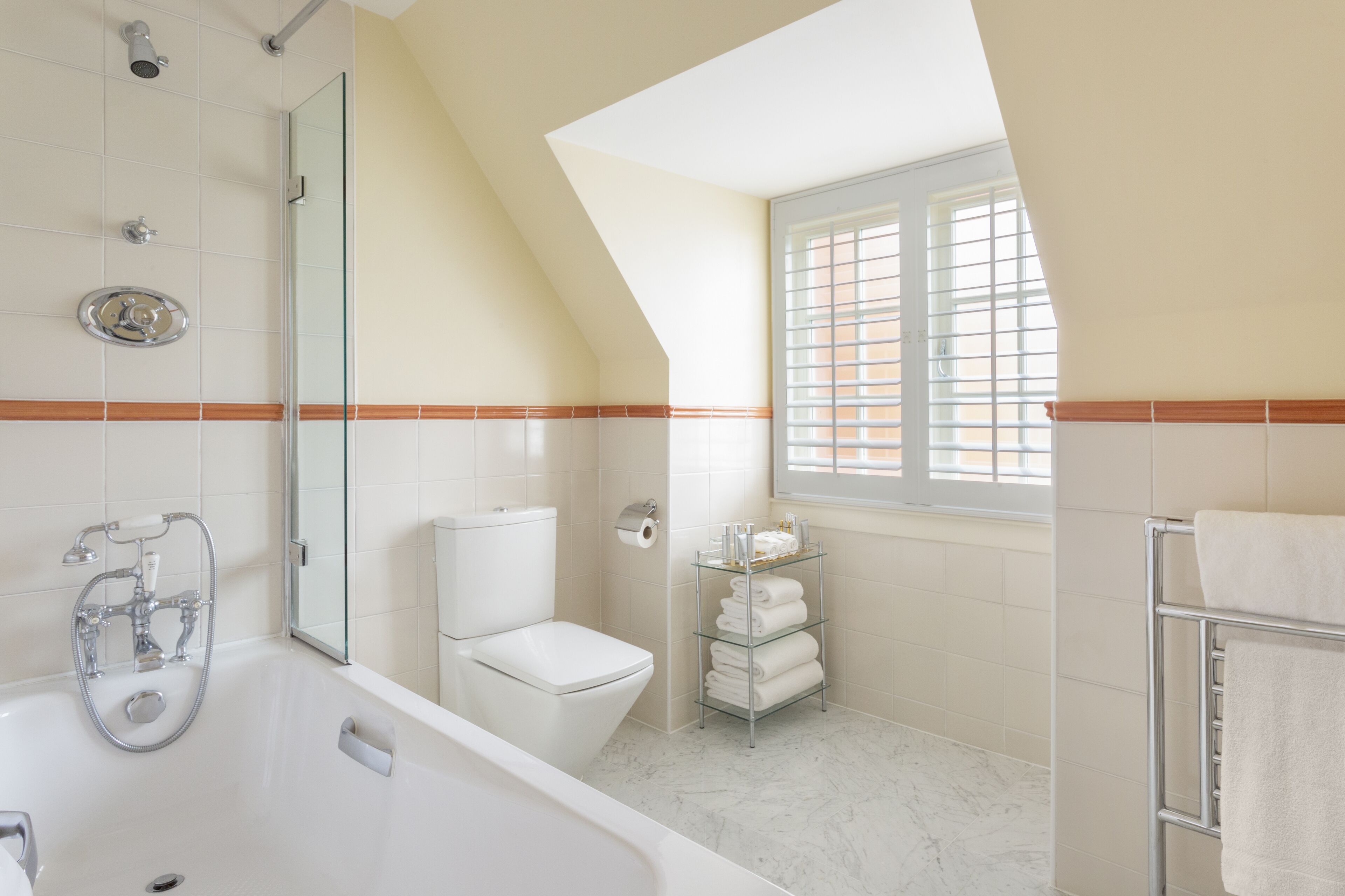A modern bathroom with a glass shower enclosure, a white toilet, a glass shelf with towels, and a window with blinds.