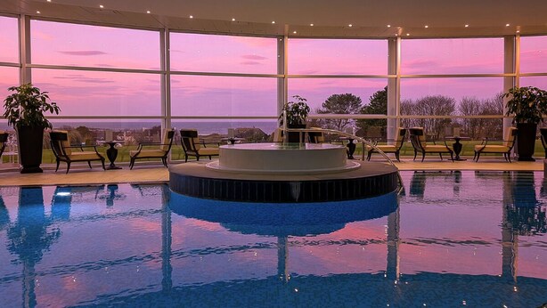 A swimming pool with a circular hot tub in the center, surrounded by lounge chairs and potted plants, with a view of the ocean through large windows. - Trump Turnberry (Girvan)