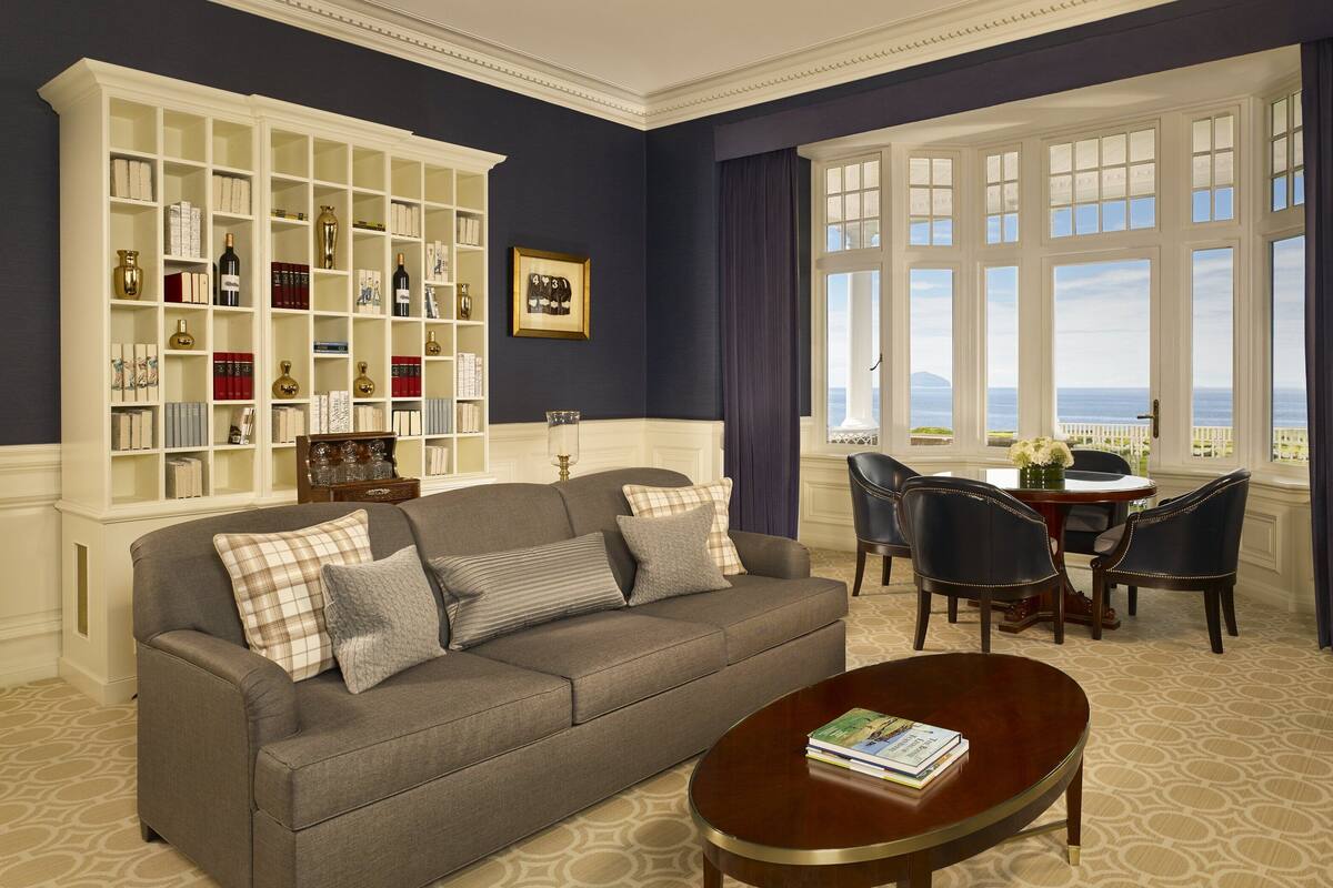 A living room with a sofa, coffee table, and dining area with chairs. There is a bookshelf with books and decorative items.