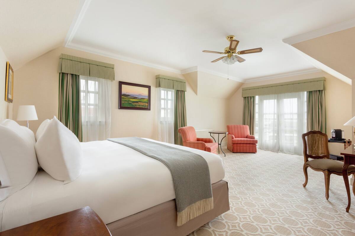 A hotel room with a large bed, a desk, two chairs, a ceiling fan, and a window with curtains.