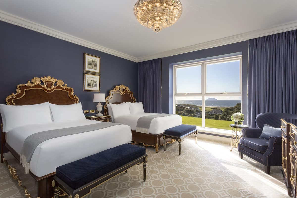 A hotel room with two large beds, a chandelier, a view of the landscape, and a side table with a lamp.