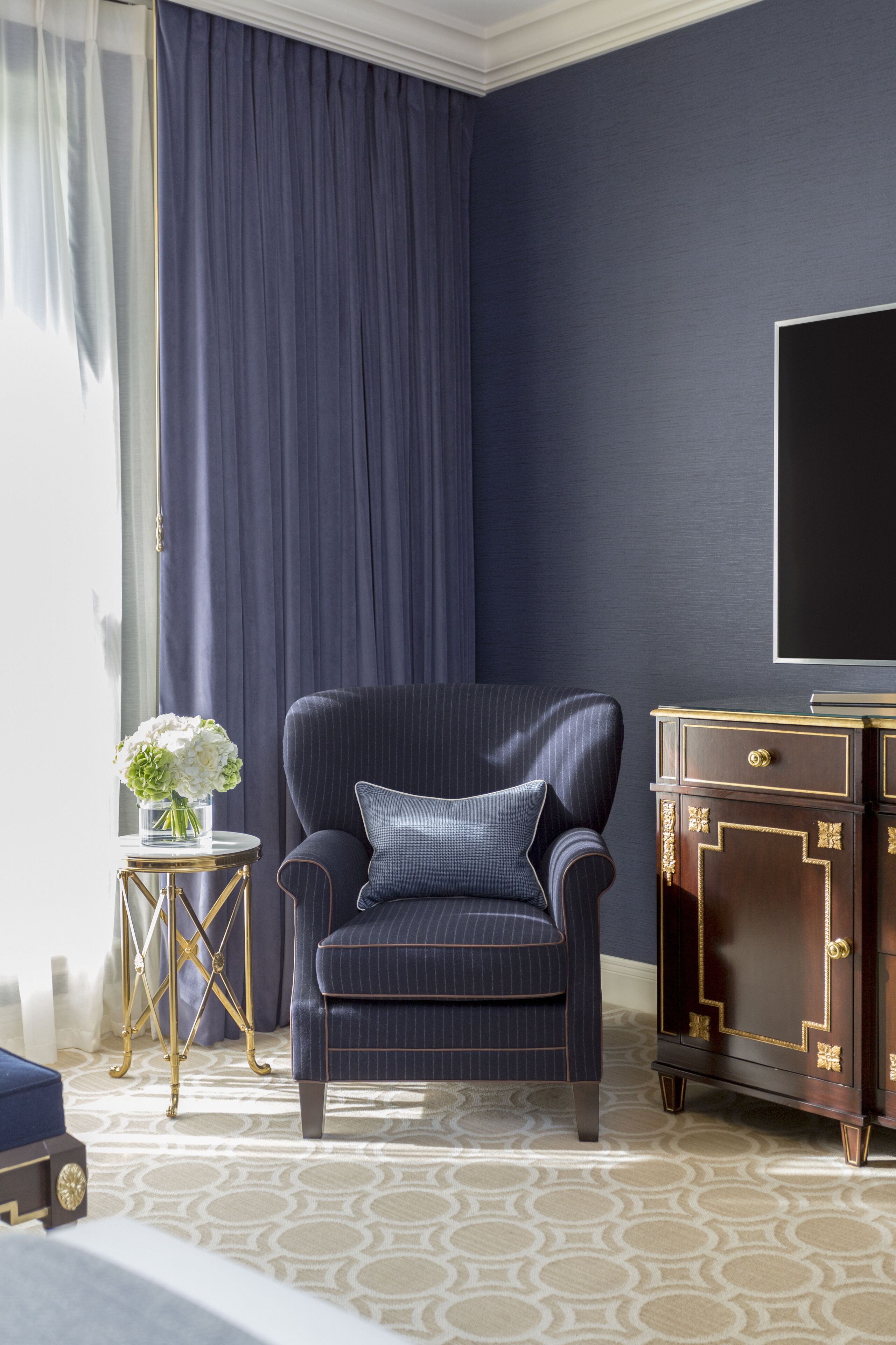 A room with a dark blue upholstered armchair, a small round side table with a vase of flowers, a dark wooden dresser with gold accents, and a large flat-screen TV mounted on the wall.