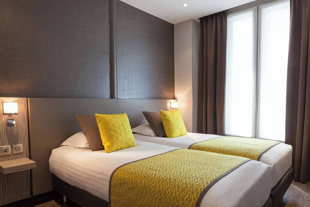 A hotel room with two beds, a headboard, bedside lamps, and a window with curtains.