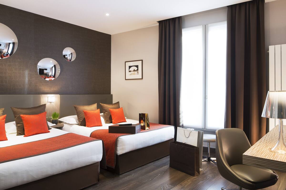 A hotel room with two beds, a desk, a chair, a lamp, and a large window with curtains.