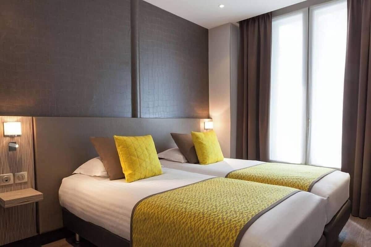 A hotel room with two beds, a nightstand, a wall-mounted lamp, and a window with curtains.