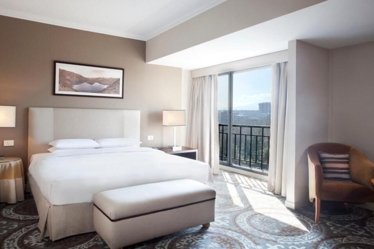 A hotel room with a large bed, a chair, a small table, a nightstand, a window with a view, and a framed picture on the wall.