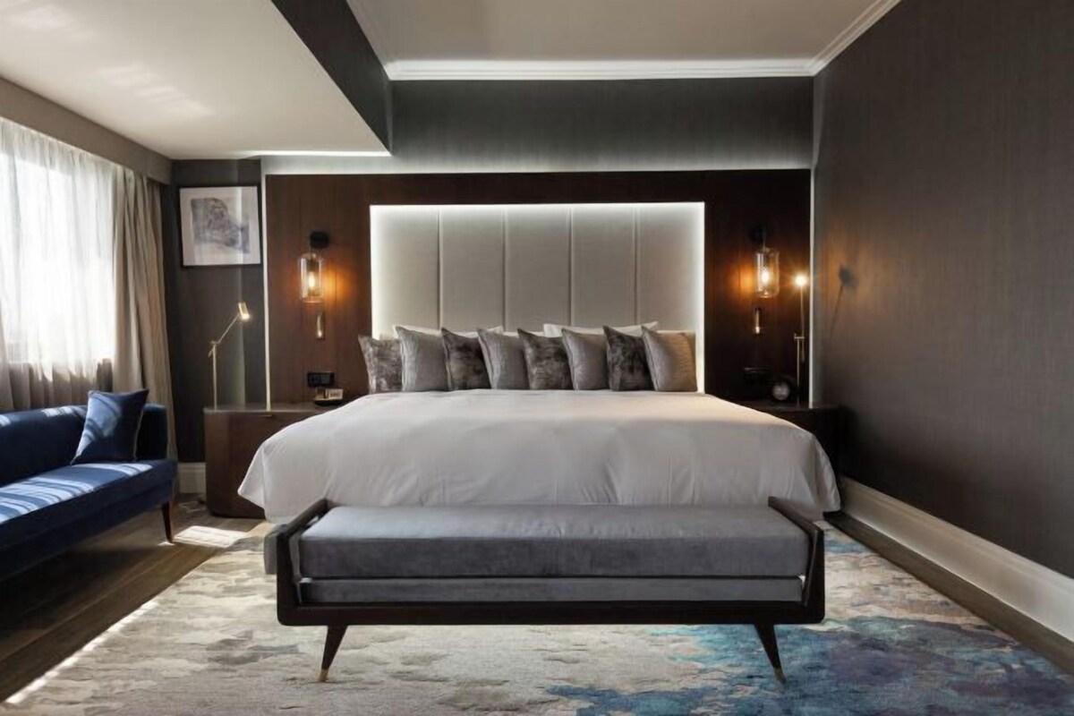 A modern bedroom with a large bed, a bench, a blue sofa, and a nightstand with a lamp.