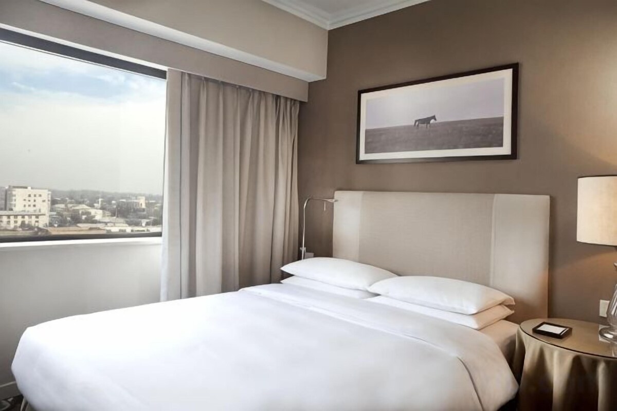 A hotel room with a large bed, a view of the city, and a framed picture on the wall.